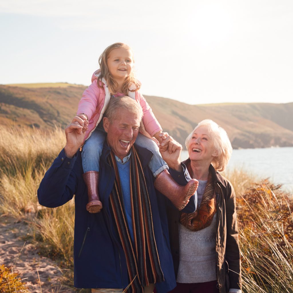 Grandfather Giving Granddaughter Ride On Shoulders As They Walk Through Sand Dunes With Grandmother